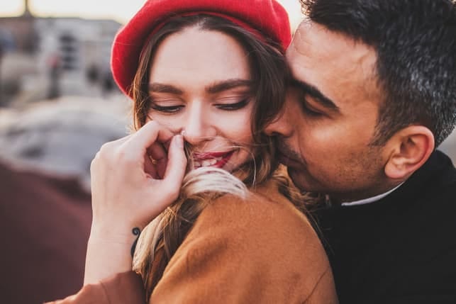 a guy is kissing his shy girlfriend in a red hat